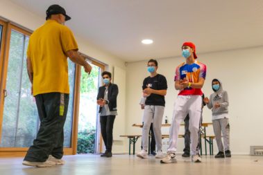 Eine Gruppe von fünf jungen Menschen tanz. Sie wird durch eine ältere Person mit gelben T-Shirt angeleitet