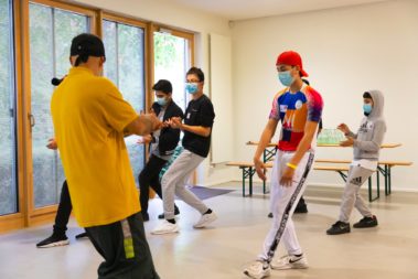 Eine Gruppe von fünf jungen Menschen tanzt. Sie wird durch eine ältere Person mit gelben T-Shirt angeleitet