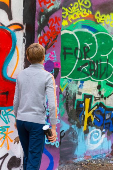 Ein junger Mensch mit kurzen Haaren, grauem Pullover mit gelben Streifen auf dem Arm und blauer Hose hät eine Sprühdose in der Hand. Er steht in die Richtung einer Wand mit vielen Graffitis