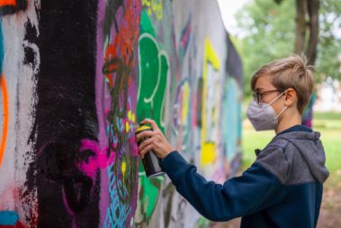 Ein junger Mensch mit kurzen Haaren, Brille, FFP2 Maske und blau-grauem Puller sprüht an eine Wand an der schon viele Graffitis zu sehen sind