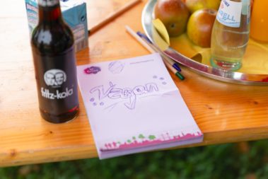 Eine Flasche Cola, ein Notizblock mit einem gezeichneten Schriftzug "vegan", mehrere Stifte, Äpfel, eine Flasche Wasser und ein Durstlöscher liegen auf einem Tisch