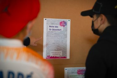 Mehrere menschen sehen sich ein Plakat an einer Pinnwand an auf dem steht: "Meine Idee für Düsseldorf" Es ist bisher nichts eingetragen