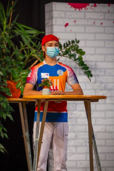 Ein junger Mensch steht an einem Stehpult. Er trägt eine rote Mütze, blaue OP-Maske, ein buntes Trikot und eine weisse Hose. Im Hintergrund sind Pflanzen zu sehen.