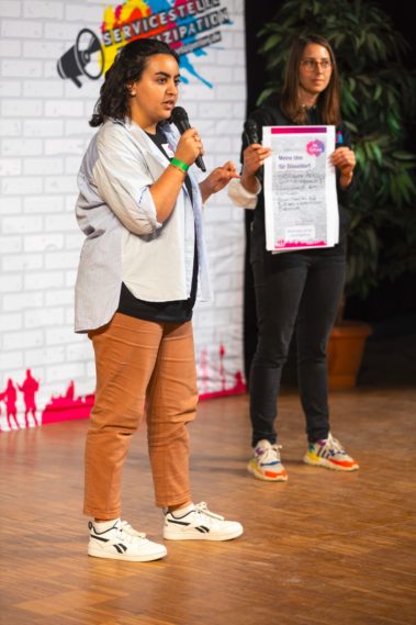 Hafida steht im Vordergrund und spricht ins Mirkofon. Rechts daneben steht Mona, sie hält ein Plakat mit Forderungen hoch.