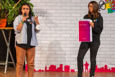 Hafida steht Links und spricht ins Mirkofon. Rechts daneben steht Mona, sie hält ein Plakat mit Forderungen hoch.